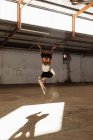 Передній вигляд молодої змішаної раси жіночої балетної танцівниці у взутті, що стрибає в повітрі у валу сонячного світла з піднятими руками під час танців у порожній кімнаті на покинутому складі — стокове фото