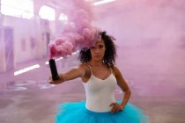 Vue de face rapprochée d'une jeune danseuse de ballet mixte portant un tutu bleu, tenant une grenade à fumée rose et regardant vers la caméra dans une pièce vide dans un entrepôt abandonné — Photo de stock