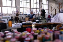 Вид сбоку на смешанную расовую женщину средних лет с помощью швейной машинки на шляпной фабрике, окруженной материалами и оборудованием, с косичками цветных нитей на переднем плане — стоковое фото