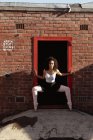 Vista frontale da vicino di una giovane ballerina di danza mista che tiene una posa da ballo in una porta in un muro di mattoni e guarda verso la telecamera, sul tetto di un edificio urbano — Foto stock
