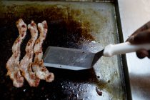 Close up of bacon frying on a hotplate and the hand of chef moving it with a spachelor in a restaurant kitchen — Stock Photo