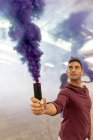 Front view close up of a young male dancer holding a purple smoke grenade and looking up at the smoke in an abandoned warehouse. — Stock Photo