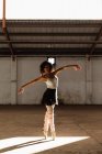 Veduta frontale di una giovane ballerina di danza mista che indossa scarpe a punta in piedi sulle dita dei piedi sotto il sole con le braccia tese mentre balla in una stanza vuota in un magazzino abbandonato — Foto stock