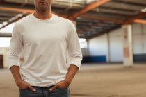 Vue de face au milieu d'un jeune homme debout les mains dans les poches dans une pièce vide dans un entrepôt abandonné — Photo de stock