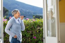Side view of a mature Caucasian woman with short grey hair standing in her garden talking on a smartphone she is holding in front of her mouth and smiling, she is seen through the open doors of her house, with a rural scene in the background — Stock Photo