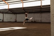 Vista lateral de una joven bailarina de ballet de raza mixta con zapatos puntiagudos saltando en el aire en el eje de la luz solar con los brazos levantados mientras baila en una habitación vacía en un almacén abandonado - foto de stock