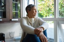 Side view close up of a mature Caucasian woman with short grey hair sitting on the counter in her kitchen looking out of the window, there are trees outside and the sun is shining — Stock Photo