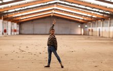 Vista frontal de um jovem dançarino de balé segurando uma pose de dança com o braço levantado em pé em um armazém abandonado olhando para a câmera — Fotografia de Stock