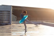 Seitenansicht einer jungen Balletttänzerin mit gemischter Rasse, die ein blaues Tutu trägt, das auf einem Bein auf ihren Zehen in einer Ballettpose steht, auf dem Dach eines städtischen Gebäudes, im Gegenlicht der Sonne — Stockfoto