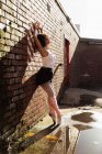 Вид збоку крупним планом молода змішана раса танцівниця балету, що стоїть на пальцях на цегляній стіні з піднятими руками на даху міської будівлі — стокове фото