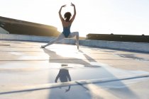 Vue arrière d'une jeune danseuse de ballet mixte debout dans une pose de ballet les bras levés, sur le toit d'un bâtiment urbain, rétro-éclairée par la lumière du soleil et reflétée dans l'eau de pluie — Photo de stock