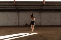 Обратный вид молодой танцовщицы балета смешанной расы в пуантах, стоящей на пальцах ног в лучах солнца с поднятыми руками, танцующей в пустой комнате на заброшенном складе — стоковое фото