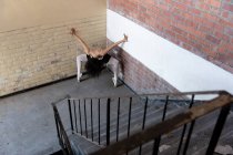 Vista frontal elevada de una joven bailarina de ballet de raza mixta que sostiene una pose de baile en los dedos de los pies con los brazos levantados y la cabeza hacia abajo en una esquina en una escalera que aterriza en un almacén abandonado - foto de stock