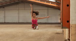Vorderansicht einer jungen gemischten Rasse Balletttänzerin, die mit ausgestreckten Armen in die Luft springt, während sie in einem leeren Raum einer verlassenen Lagerhalle tanzt — Stockfoto