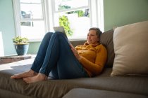 Вид сбоку на взрослую белую женщину с короткими седыми волосами, сидящую на диване в гостиной, улыбающуюся и пользующуюся планшетным компьютером, солнечное окно на заднем плане — стоковое фото