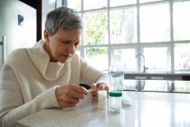 Vue de côté gros plan d'une femme caucasienne mature avec les cheveux gris courts assis dans sa cuisine regardant ses médicaments, avec des bouteilles de pilules, une boîte de pilules hebdomadaire et un verre d'eau sur le comptoir à côté d'elle — Photo de stock