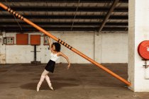 Vista lateral de uma jovem mista dançarina de balé usando sapatos pontiagudos dançando nos dedos dos pés segurando um poste estrutural em uma sala vazia em um armazém abandonado — Fotografia de Stock