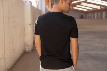 Rückseite Nahaufnahme eines jungen Mannes in einem schwarzen T-Shirt, der mit zur Seite gedrehtem Kopf in einer verlassenen Lagerhalle steht — Stockfoto