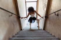 Vue de face surélevée d'une jeune danseuse de ballet mixte tenant les mains courantes et dansant au bas d'un escalier dans un entrepôt abandonné — Photo de stock