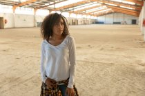 Front view of a young mixed race woman with shoulder length curly hair standing and looking away in an abandoned warehouse — Stock Photo