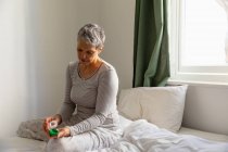 Nahaufnahme einer reifen kaukasischen Frau mit kurzen grauen Haaren, die auf ihrem Bett sitzt und zu Hause Medikamente einnimmt — Stockfoto