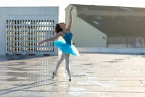 Veduta laterale di una giovane ballerina di danza mista che indossa un tutù blu in piedi in una posa di balletto con la testa indietro e un braccio alzato, sul tetto di un edificio urbano — Foto stock