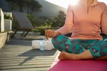 Vorderseite Unterteil einer Frau in Sportkleidung, die in Yogaposition auf einer Matte sitzt und in ihrem Garten meditiert, im Gegenlicht des Sonnenlichts mit ländlichem Blick im Hintergrund — Stockfoto