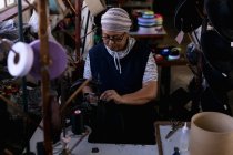 Підвищений вид спереду жінки середнього віку змішаної раси з використанням швейної машини на капелюшній фабриці, оточеної матеріалами — стокове фото