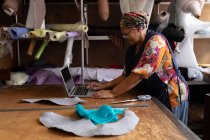 Seitenansicht einer Frau mittleren Alters, die an einem Tisch steht und einen Laptop benutzt, während sie in einer Hutfabrik arbeitet. — Stockfoto