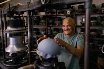 Portrait of a smiling senior mixed race man holding his hands above the top of a hat that has been shaped on a piece of equipment in the workshop at a hat factory — Stock Photo