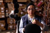 Front view close up of a middle aged mixed race woman holding and adding the finishing touches to a small hat in the workshop at a hat factory, with boxes of materials in the background — Stock Photo