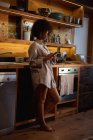 Vista lateral de cerca de una joven mujer de raza mixta con una camisa de pie usando un teléfono inteligente en casa en su cocina - foto de stock