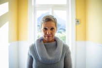 Portrait of a mature Caucasian woman with short grey hair wearing a cowl neck sweater, standing in front of a window at home looking straight to camera and smiling, with a rural view in the background — Stock Photo