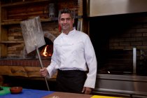 Retrato close-up de um cozinheiro masculino caucasiano de meia idade sorridente segurando uma casca de pizza e em pé junto a um forno de pizza em uma cozinha do restaurante — Fotografia de Stock
