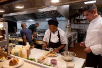 Vista laterale da vicino di un cuoco caucasico di mezza età che supervisiona una giovane cuoca afroamericana che prepara un piatto in una cucina da ristorante, con il personale della cucina impegnato a lavorare sullo sfondo — Foto stock
