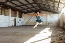 Передній вигляд молодої змішаної раси танцівниці балету, що стрибає в повітрі з простягнутими руками під час танців у порожній кімнаті на покинутому складі — стокове фото