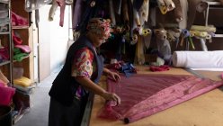 Vista laterale di una donna di razza mista di mezza età in piedi a un tavolo che lavora con tessuto in una fabbrica di cappelli . — Foto stock