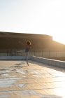 Вид сзади на молодую балетную танцовщицу смешанной расы, стоящую на одной ноге, с другой поднятой ногой и поднятыми руками, на крыше городского здания, освещенного солнечным светом — стоковое фото