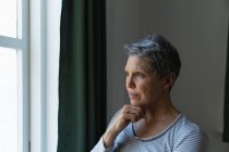 Vista lateral de cerca de una mujer caucásica madura con el pelo gris corto de pie y mirando por la ventana en casa con la mano debajo de la barbilla - foto de stock