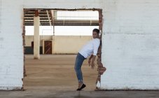 Vista lateral de um jovem bailarino vestindo jeans posando em uma porta em um quarto vazio em um armazém abandonado — Fotografia de Stock