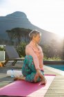 Vue de côté gros plan d'une femme blanche mature aux cheveux gris court portant des vêtements de sport assis sur un tapis dans une position de yoga, s'exerçant près de la piscine dans son jardin, avec une vue rurale en arrière-plan — Photo de stock