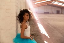 Vue de côté gros plan d'une jeune danseuse de ballet mixte portant un tutu bleu debout contre un mur dans une pièce vide dans un entrepôt abandonné, un rayon de soleil devant elle — Photo de stock