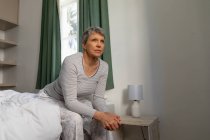 Vue de côté gros plan d'une femme blanche mature aux cheveux gris courts assise sur le côté de son lit à la maison les mains jointes, regardant ailleurs — Photo de stock