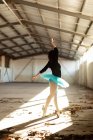 Вид сбоку на молодую балетную танцовщицу смешанной расы в синей пачке и пуантах, танцующую на пальцах ног в лучах солнца в пустой комнате на заброшенном складе — стоковое фото