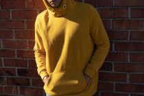 Vista frontale sezione centrale di un giovane uomo che indossa una felpa gialla con cappuccio appoggiata a un muro di mattoni con le mani nella tasca anteriore — Foto stock
