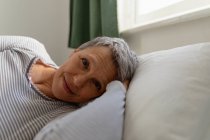Vista frontal de cerca de una mujer caucásica madura con el pelo gris corto acostada en su lado en la cama en casa, mirando a la cámara y sonriendo - foto de stock