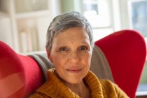 Portrait gros plan d'une femme blanche mature aux cheveux gris court assise dans un fauteuil rouge dans son salon regardant vers la caméra et souriant légèrement, une fenêtre ensoleillée en arrière-plan — Photo de stock
