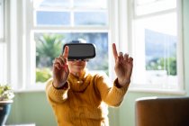 Front view close up of a mature Caucasian woman with short grey hair sitting at home in her living room wearing a VR headset with her arms outstretched in from of her and her fingers pointing up, back lit by sunlight from a window behind her — Stock Photo