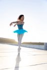 Вид спереди на молодую балетную танцовщицу смешанной расы в голубой пачке и пуантах, танцующую на крыше городского здания, освещенную солнечным светом — стоковое фото
