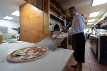 Vue latérale de près d'un chef caucasien d'âge moyen ramassant une pizza avec une peau de pizza pour la mettre au four dans une cuisine de restaurant — Photo de stock